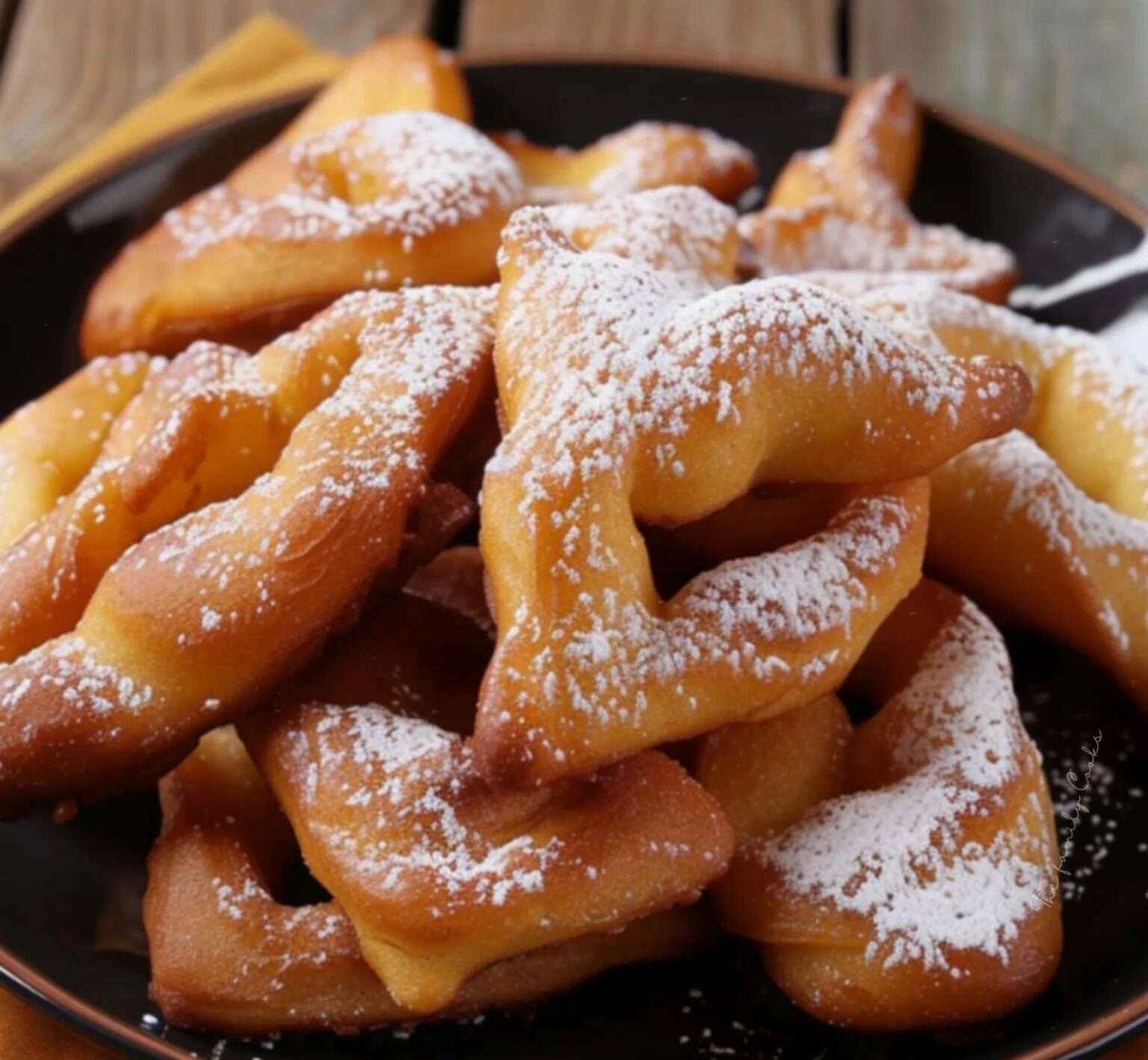 Bugnes dorées et torsadées, saupoudrées de sucre glace, présentées sur une assiette rustique avec une tasse de thé et des tranches de citron en arrière-plan.