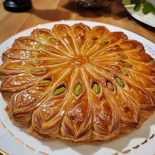 Une galette des rois dorée, garnie de crème de pistache onctueuse, décorée de pistaches concassées et saupoudrée de sucre glace.