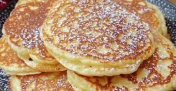 Pile de pancakes dorés au yaourt sur une assiette avec fruits rouges et sirop d’érable, posée sur une table en bois rustique.