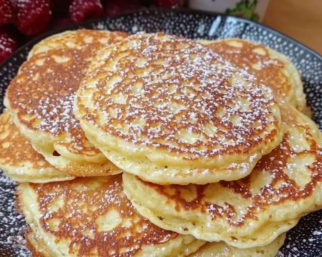 Pile de pancakes dorés au yaourt sur une assiette avec fruits rouges et sirop d’érable, posée sur une table en bois rustique.