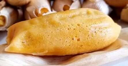 Tamales dulces de piña abiertos en hoja de maíz, mostrando su interior amarillo y trozos de piña, sobre una mesa rústica con taza de chocolate caliente.