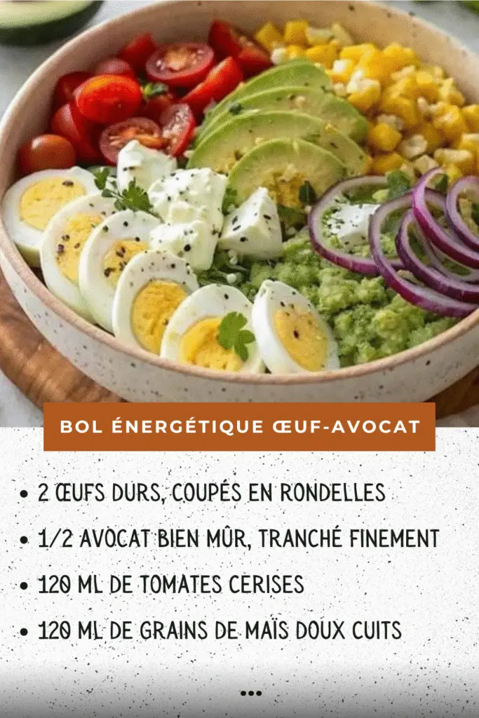 Bol énergétique composé de tranches d’œufs durs, avocat, tomates cerises, maïs, oignon rouge et feta, présenté sur une table en bois avec herbes fraîches.