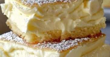 Gros plan sur deux carrés à la crème vanille empilés sur une assiette blanche. Chaque carré est composé de deux couches de génoise dorée garnies d'une crème vanille onctueuse, saupoudré de sucre glace et surmonté d’une rosace de chantilly.