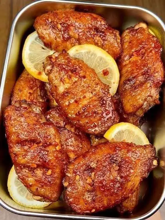 Ailes de poulet dorées nappées de sauce miel-citron, servies chaudes, parsemées de sésame et d’oignons verts sur plat blanc.