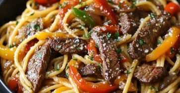 Assiette de pâtes penne avec lanières de steak grillé, poivrons rouges et verts, oignons et sauce crémeuse, parsemée de parmesan et de persil frais.