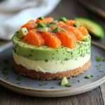 Part de cheesecake salé au saumon fumé, avocat et ciboulette, servie sur une assiette blanche avec zeste de citron.