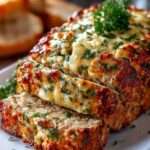 Pain de viande au poulet, ail et parmesan doré au four, tranché sur une planche, accompagné d’herbes fraîches et de légumes rôtis.