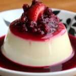 Panna cotta légère à la vanille servie dans une verrine, nappée d’un coulis de fruits rouges brillant et décorée de framboises fraîches et de menthe.