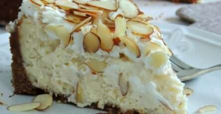 Cheesecake à l’amande et à l’Amaretto, surmonté d’amandes grillées dorées, sur fond de table en bois clair, tranché pour révéler sa texture onctueuse et dorée.
