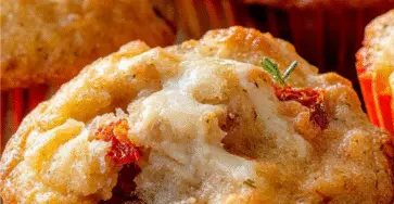 Muffins salés dorés au fromage de chèvre et tomates séchées, garnis d’herbes de Provence et servis sur une planche en bois pour l’apéritif.