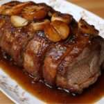 Rôti de porc caramélisé au miel tranché, nappé de sauce brillante miel-soja avec échalotes et romarin, servi dans une cocotte avec garniture.