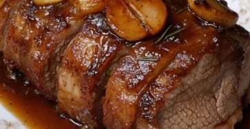 Rôti de porc caramélisé au miel tranché, nappé de sauce brillante miel-soja avec échalotes et romarin, servi dans une cocotte avec garniture.