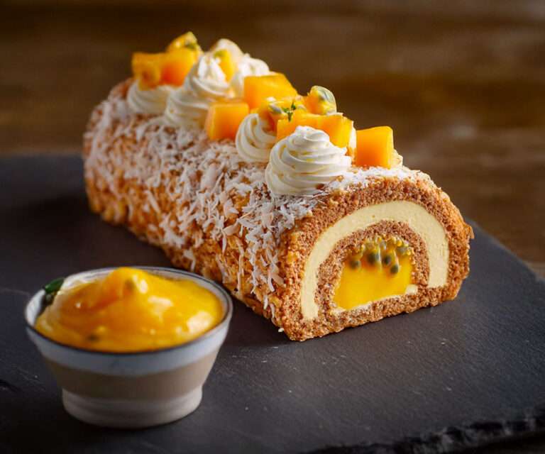 Bûche de Noël mangue passion coco glacée, décorée de dés de mangue fraîche et copeaux de coco, présentée sur un plat de fête tropical.