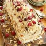 Bûche roulée garnie de crème au mascarpone et framboises, décorée d’amandes effilées, fruits secs et sucre glace sur un plat de fête.