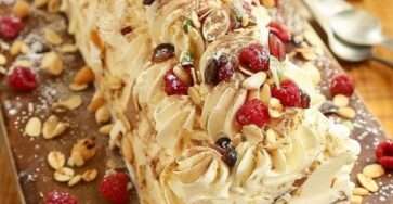 Bûche roulée garnie de crème au mascarpone et framboises, décorée d’amandes effilées, fruits secs et sucre glace sur un plat de fête.