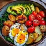 Bowl de légumes rôtis coloré, pommes de terre dorées, œufs mollets, avocat tranché et tomates cerises sur lit de verdure fraîche.