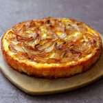 Gâteau salé moelleux aux oignons dorés et fromage fondu, coupé en parts, mie fondante, servi tiède sur une planche avec salade.