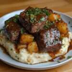 Assiette de bœuf fondant mijoté dans une sauce brune onctueuse, servi avec une purée maison crémeuse et des cubes de pommes de terre, parsemé de persil frais.