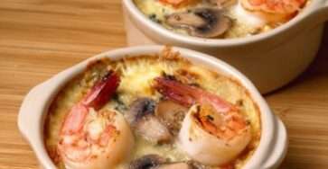 Cassolette de Saint-Jacques, crevettes et champignons dans une sauce crémeuse gratinée, servie bien chaude avec persil frais et croûte dorée.
