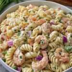 Salade de pâtes froides aux crevettes, petits pois, carottes râpées et oignon rouge, enrobée d’une sauce crémeuse, servie dans un grand saladier.