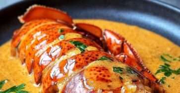 Queue de homard nappée de sauce crémeuse, chair tendre légèrement dorée, présentée sur une assiette élégante avec persil frais.