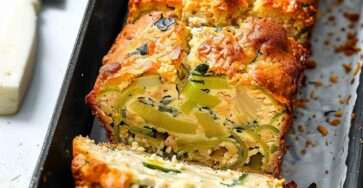 Tranches de cake salé aux poireaux et fromage de chèvre, texture moelleuse avec morceaux fondants, servies sur une planche avec herbes fraîches.