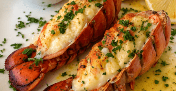 Queues de homard grillées au four, chair dorée et nappée de beurre citronné à l’ail, servies avec du persil frais et des quartiers de citron.