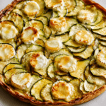 Tarte fine aux courgettes, chèvre et miel sur pâte feuilletée dorée, garnie de rondelles de courgettes, fromage de chèvre fondu et filet de miel.