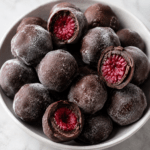 Bouchées glacées aux framboises enrobées de chocolat noir, avec une coque croquante et un cœur fruité, servies sur du papier cuisson.