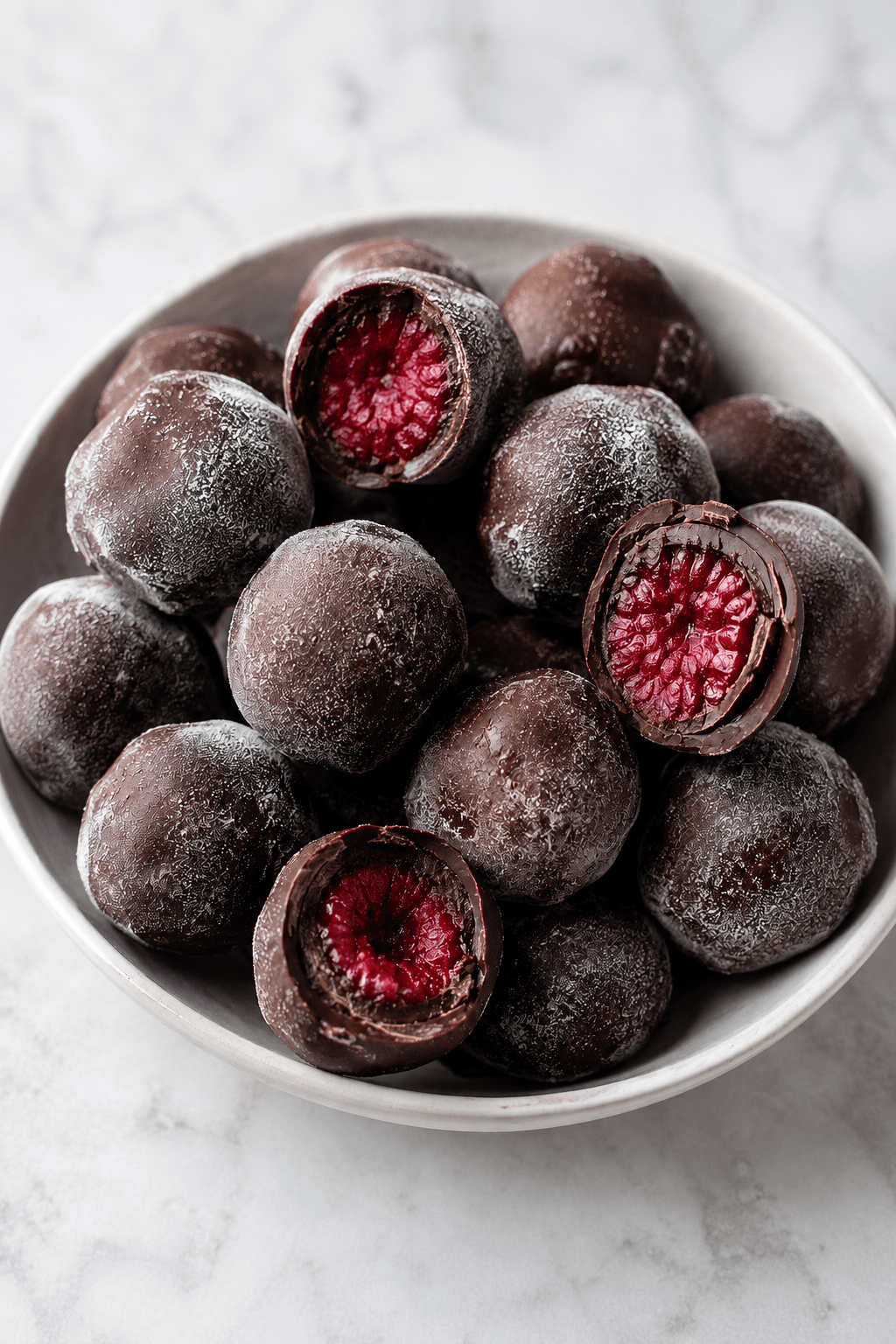 Bouchées glacées aux framboises enrobées de chocolat noir, avec une coque croquante et un cœur fruité, servies sur du papier cuisson.