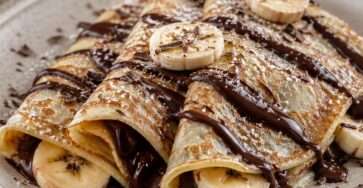 Crêpes fines et moelleuses garnies de chocolat fondant, de rondelles de banane fraîche et saupoudrées de sucre glace dans une assiette