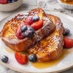Tranches de pain perdu dorées et moelleuses servies avec des fruits rouges frais, un filet de miel et une légère touche de sucre glace.