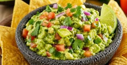 Guacamole maison frais et crémeux servi dans un bol, avec morceaux de tomate, oignon rouge, coriandre et chips tortilla autour.