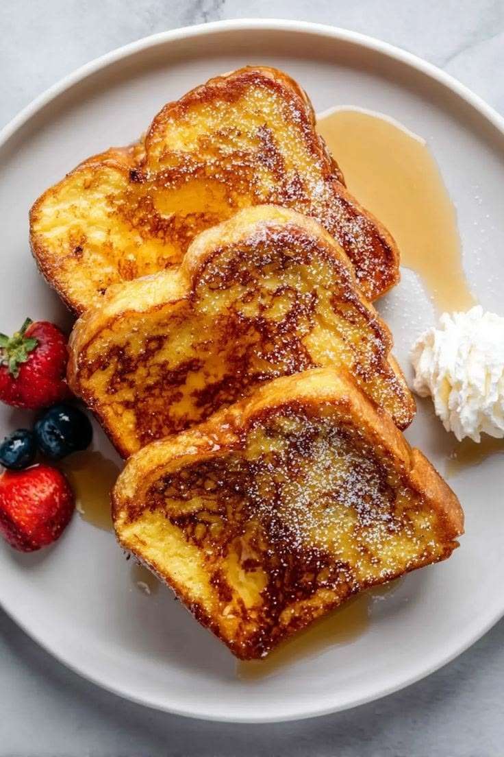 Tranches de pain perdu moelleux et doré, nappées de sirop d’érable, saupoudrées de sucre glace et servies avec fraises et myrtilles.