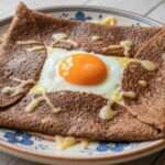 Galette complète bretonne au sarrasin garnie de jambon blanc, fromage râpé fondu et œuf au jaune coulant, pliée en carré.