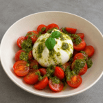 Salade fraîche de burrata crémeuse servie avec tomates cerises coupées, pesto vert, huile d’olive et feuilles de basilic dans une assiette creuse.