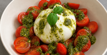 Salade fraîche de burrata crémeuse servie avec tomates cerises coupées, pesto vert, huile d’olive et feuilles de basilic dans une assiette creuse.