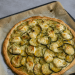 Tarte fine dorée aux courgettes en rondelles, fromage de chèvre fondant, miel brillant, huile d’olive et herbes de Provence sur pâte feuilletée.