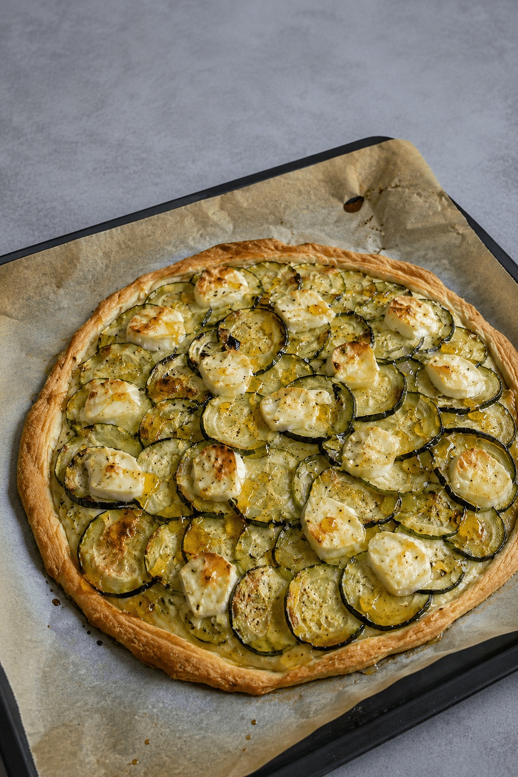 Tarte fine dorée aux courgettes en rondelles, fromage de chèvre fondant, miel brillant, huile d’olive et herbes de Provence sur pâte feuilletée.