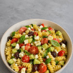 Salade de pâtes méditerranéenne colorée avec tomates cerises, concombre, feta en morceaux, olives noires, basilic frais et huile d’olive.