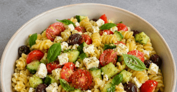 Salade de pâtes méditerranéenne colorée avec tomates cerises, concombre, feta en morceaux, olives noires, basilic frais et huile d’olive.