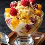Coupe glacée tropicale servie dans un verre avec boules de glace vanille et mangue, dés de mangue fraîche, framboises, coulis fruité et amandes effilées grillées.