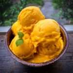 Boules de glace à la mangue maison servies dans une coupe, avec une texture crémeuse jaune doré et une feuille de menthe fraîche en décoration.