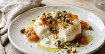 Burrata ouverte à la confiture d’abricots et aux pistaches concassées, servie avec du pain grillé dans une assiette claire.