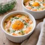 Quatre ramequins d’œufs cocotte au saumon fumé, à la crème et au fromage, cuits au bain-marie et garnis d’aneth frais.