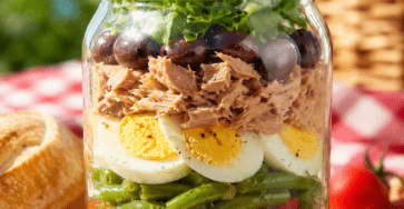 Salade niçoise présentée dans un grand bocal en verre transparent, avec vinaigrette au fond, haricots verts, tomates, pommes de terre, thon, œufs durs, olives noires et salade verte disposés en couches colorées.
