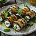 Roulés d’aubergine grillée garnis de ricotta crémeuse et de menthe fraîche, disposés sur une assiette en céramique avec de l’huile d’olive et du citron.