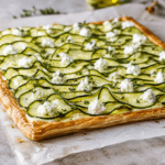 Tarte fine rectangulaire en pâte feuilletée garnie de ricotta et de fines rondelles de courgettes disposées en écailles, dorées sur les bords et parfumées aux herbes.